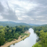 A wide river curves through dense green forest under a cloudy sky, with distant hills visible on the horizon and a sandy riverbank along one side.