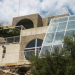 Modern concrete building with large arched windows and a slanted glass structure, surrounded by greenery, under a partly cloudy sky. White railings border the terraces and balconies.