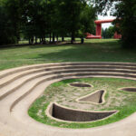 A sunken, concrete amphitheater with steps forms the shape of a smiling face, set in a grassy park with trees and a large red sculpture in the background.