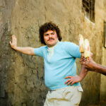 A man with curly hair and a mustache, wearing a light blue shirt and beige pants, leans against a textured wall. Another person holds two ears of corn on sticks toward him.