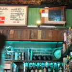 A bar interior with a menu board listing cocktails, a shelf with bottles and mugs, neon lights, and a framed painting of a nighttime bridge scene above wooden paneling.