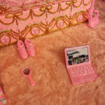 A pink laptop, a pair of pink shoes, and a pink hand mirror rest on a fluffy pink rug next to an ornate, pink-and-gold decorated bed. The scene has a whimsical, highly decorative, and monochromatic pink theme.
