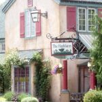 A rustic building with red shutters and a sign reading The Herbfarm hangs by the entrance. The exterior is surrounded by greenery, flowers, and a small garden.