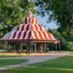 A striped red-and-white pavilion with a domed roof stands in a park surrounded by green grass and trees. Picnic tables are underneath, and sunlight filters through the scene.
