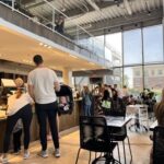 A modern, busy café with large windows and high ceilings. People are ordering at the counter, sitting at tables, and a man is holding a baby in a carrier while standing in line. Natural light fills the space.