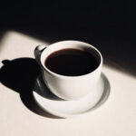A white ceramic cup filled with dark coffee sits on a matching saucer, casting a strong shadow on a sunlit white surface.