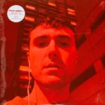 A person with short hair wearing earphones takes a close-up selfie at night in front of a tall building, with a strong red filter over the entire image. A circular white sticker in the top left corner displays text.