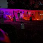 A house decorated for Halloween at night with purple and orange lights, fake gravestones in the yard, hanging skeletons, and a large inflatable pumpkin near the garage. The scene is dark and spooky.