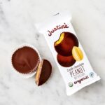 A package of Justins organic milk chocolate peanut butter cups on a marble surface, with one whole cup and one half cup next to the opened wrapper.