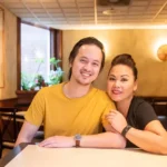 A smiling man in a yellow shirt and a woman in a black shirt sit closely together at a restaurant table, with warm lighting and an inviting, cozy atmosphere.