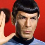 A man with pointed ears and straight-cut bangs raises his right hand in a Vulcan salute against a red background, wearing a light blue uniform.