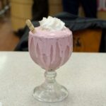A pink milkshake in a glass goblet, topped with whipped cream and a wafer stick. The milkshake is overflowing and dripping down the sides of the glass, set on a white table.