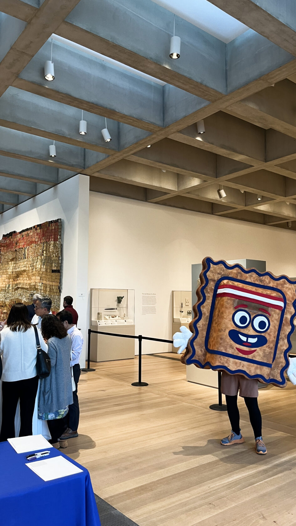 A person in a smiling pastry costume with arms and legs stands in an art museum, while visitors gather near exhibits and art displays in the background. Natural light filters through the ceiling.