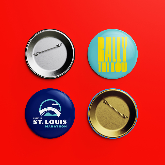 Four round buttons on a red background; the top left and bottom right show the metal backside, while the top right says Rally The Lou in yellow, and the bottom left shows a blue Greater St. Louis Marathon logo.