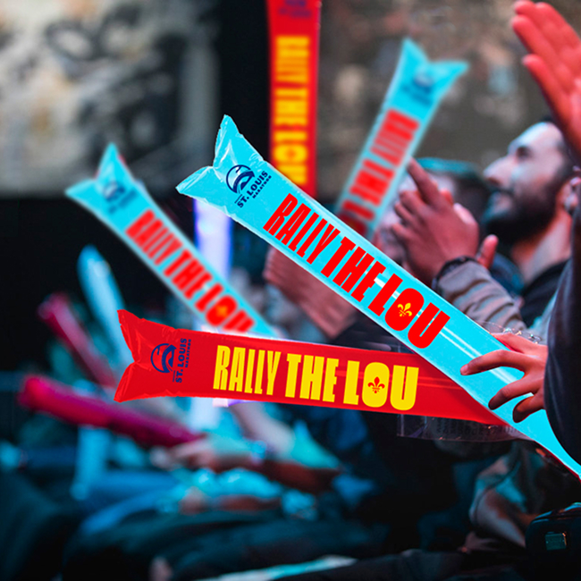 A row of people clapping and holding inflatable cheering sticks that read Rally The Lou in red and blue at an indoor event.