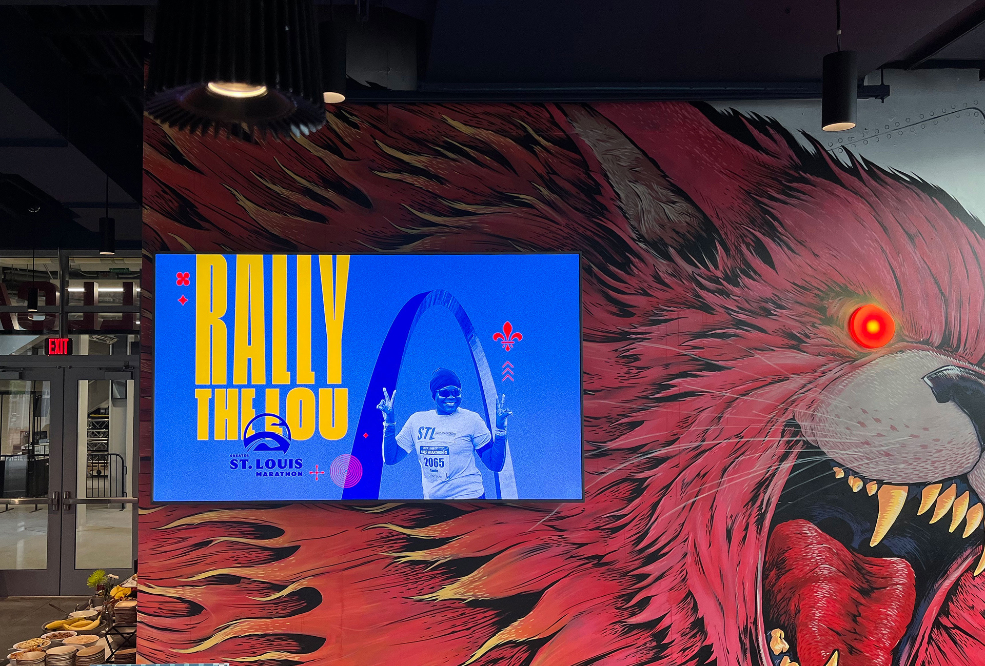A brightly colored mural of a roaring red wolf with a glowing eye is on a wall. A TV screen shows “Rally the Lou” and a person in a St. Louis t-shirt, with blue and yellow graphics and city branding.
