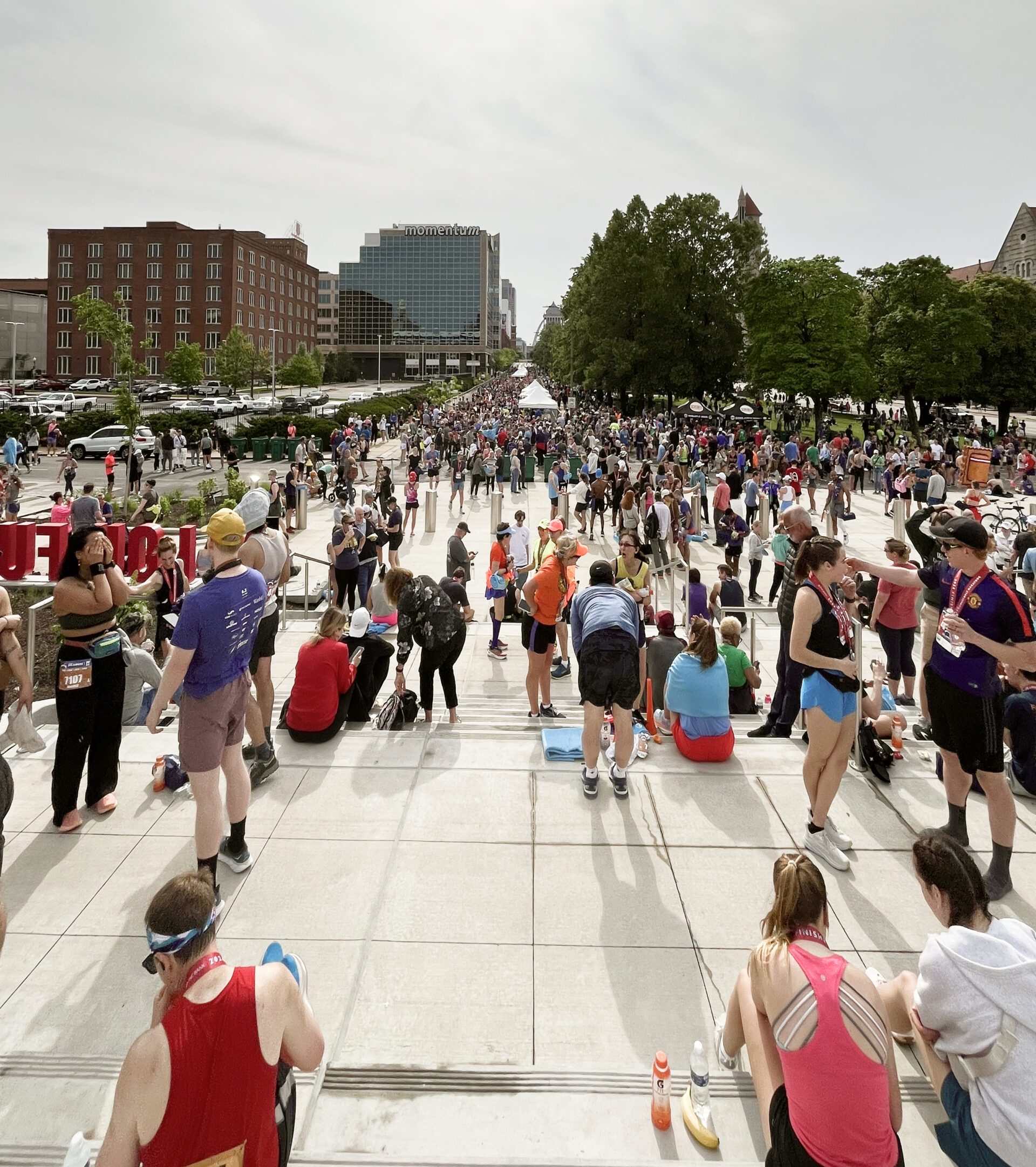 A large crowd of people gathers outside on wide steps and a plaza during a sunny day, many wearing athletic clothing and race bibs, suggesting a running event or marathon. Buildings and trees line the background.