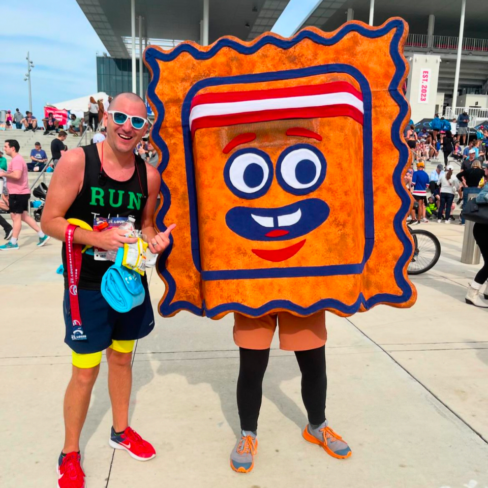 A smiling runner poses next to a person in a large, square, cartoon cracker costume with a face, at an outdoor event with crowds and modern buildings in the background.