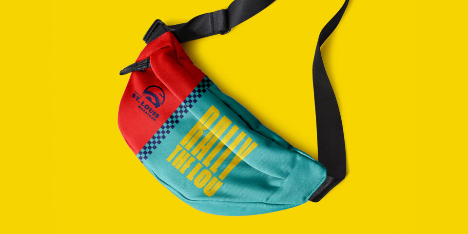 A colorful crossbody bag with a black strap lies on a yellow background. The bag is red and teal with the text RALLY THE LOU and ST. LOUIS CITY SC next to a logo and a blue checkered stripe.