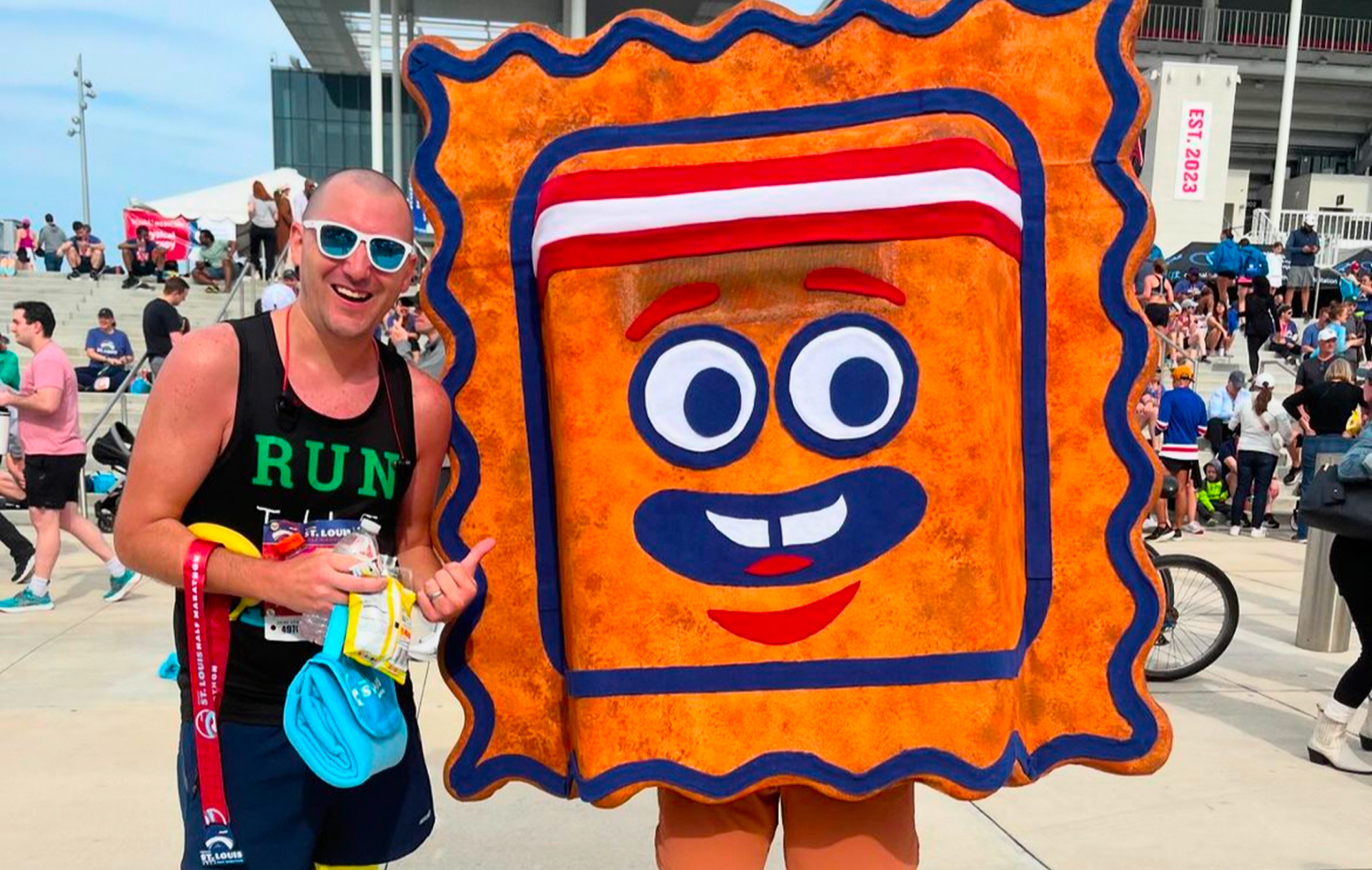 A smiling runner wearing sunglasses poses next to a person in a large, colorful cartoon ravioli costume at an outdoor event with people in the background.