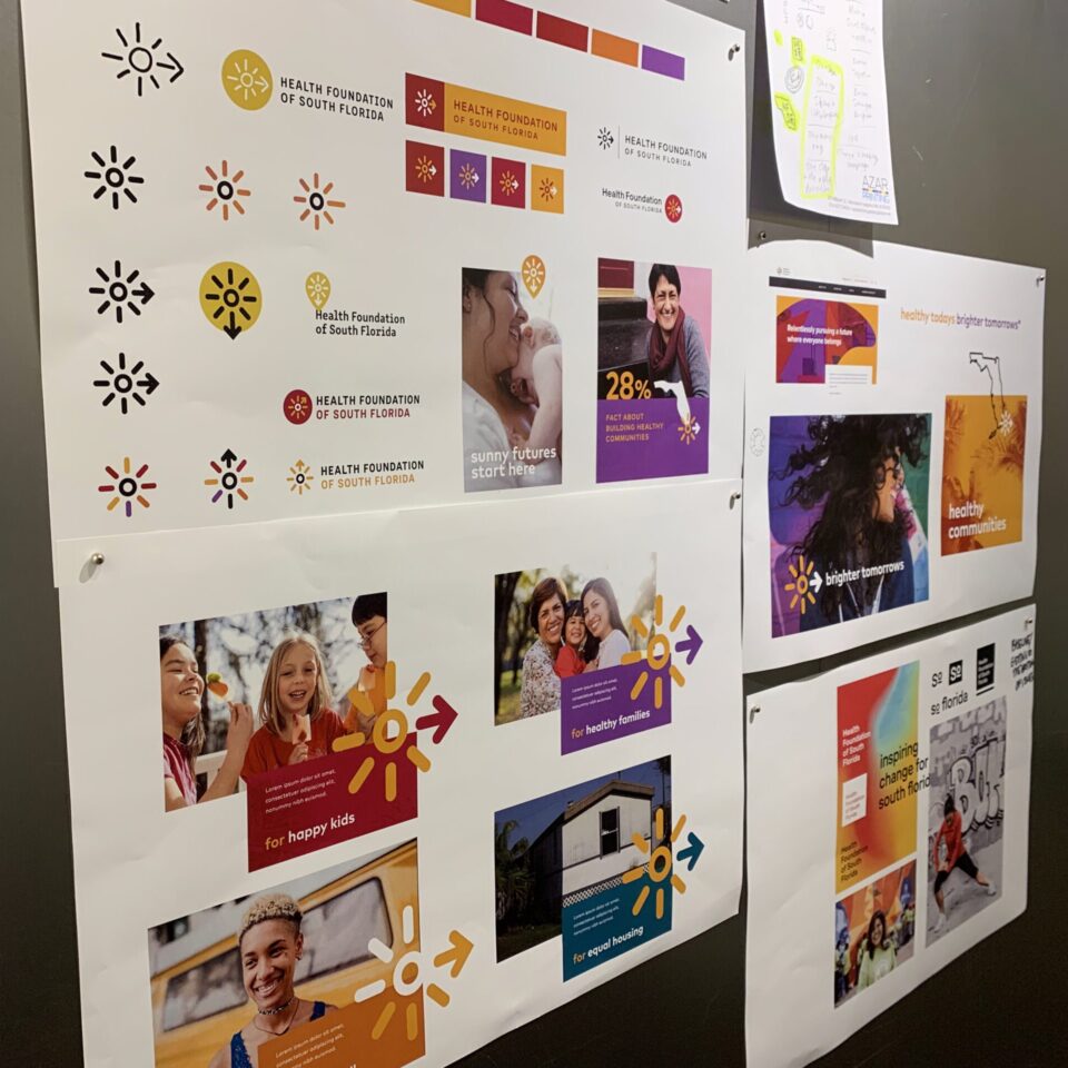 Printed branding materials and logo designs for the Health Foundation of South Florida are displayed on a wall, showing colorful logos, sample brochures, color palettes, and images of diverse, smiling people.