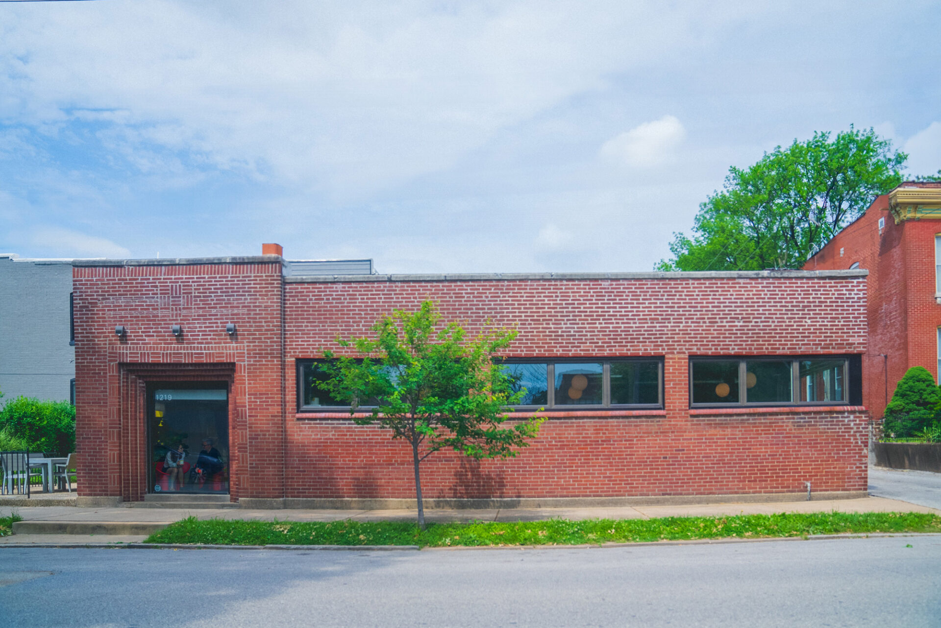 A single-story red brick building with a row of windows, a small tree in front, and a sidewalk along the street under a partly cloudy sky.