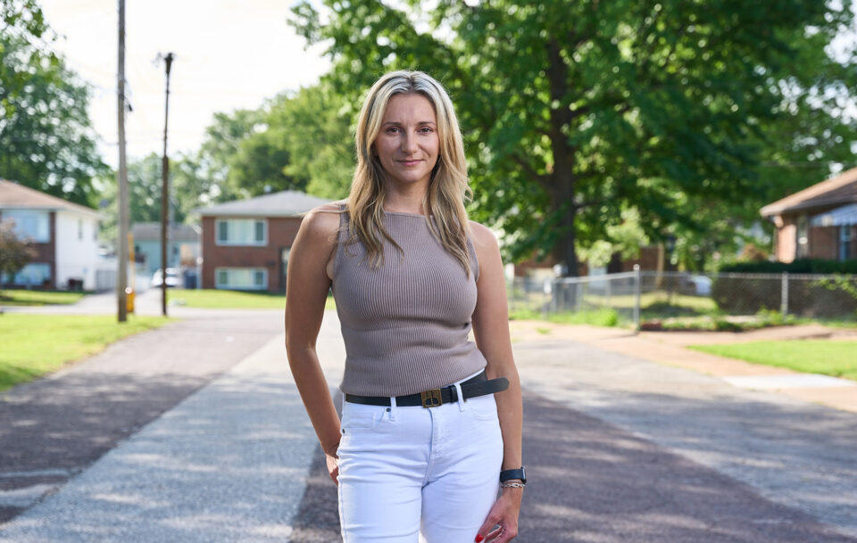 A woman with long blonde hair, wearing a sleeveless taupe top and white pants, stands confidently on a quiet residential street lined with houses and trees on a sunny day.