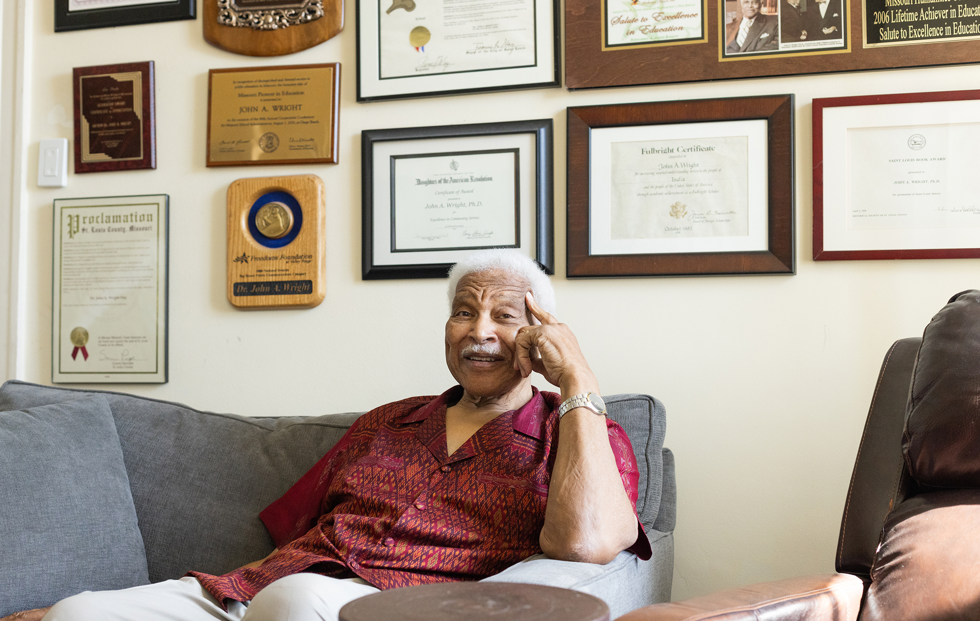 An older man with gray hair sits on a sofa, smiling and pointing to his temple. He is wearing a red patterned shirt, and behind him, a wall is filled with framed certificates, awards, and proclamations.