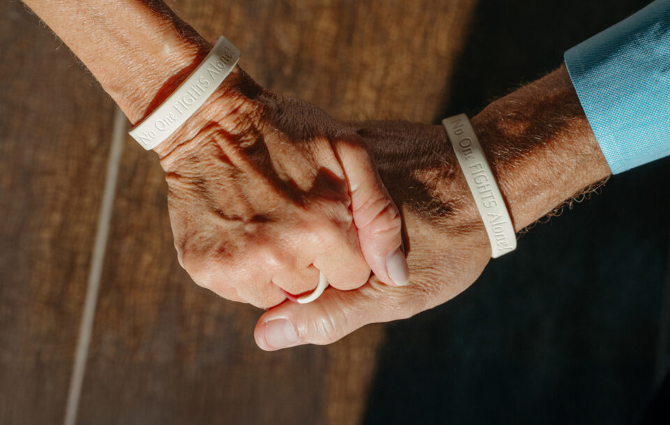 Two people hold hands, each wearing a white wristband that reads No One Fights Alone. The scene is warmly lit, emphasizing the support and connection between them.