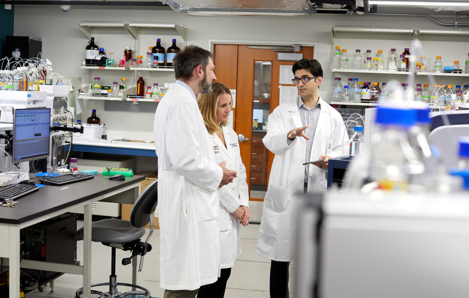 Three scientists in white lab coats stand and talk in a modern laboratory filled with equipment, shelves of bottles, and computers. One person gestures while the others listen attentively.
