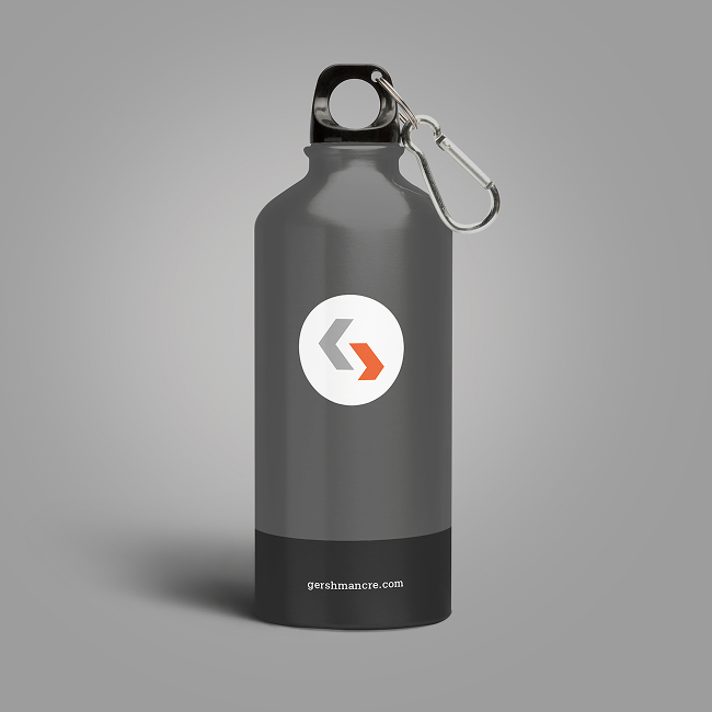 A gray metal water bottle with a black base, a silver carabiner attached to the lid, and a circular logo featuring two arrows—one gray, one orange—on the front. The website gershimacre.com appears near the base.