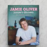 A cookbook titled Jamie Oliver: Jamies Dinners – the essential cookbook lies on a marble surface, featuring a photo of Jamie Oliver sitting casually in a white t-shirt and jeans.