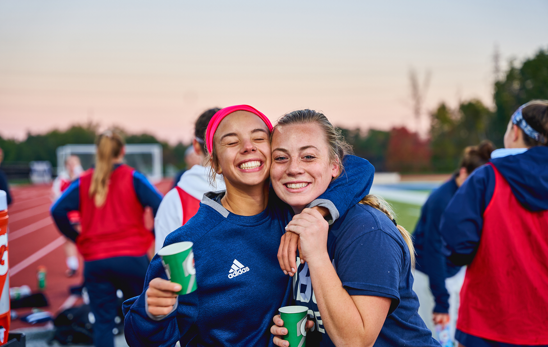 Two young women in sportswear hug and smile at the camera while holding green paper cups on an outdoor track, with other teammates in the background. The sky is pink, indicating either sunrise or sunset.
