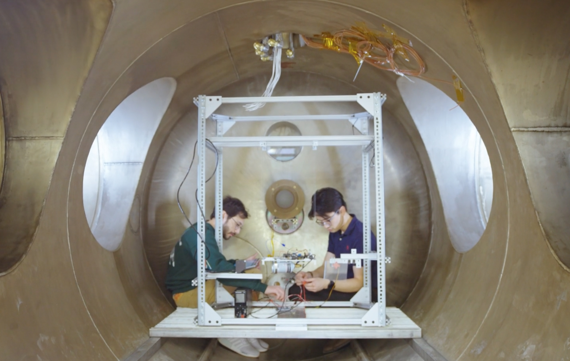 Two people work inside a large metallic cylindrical chamber, assembling electronic equipment on a white metal frame, surrounded by wires and scientific instruments.