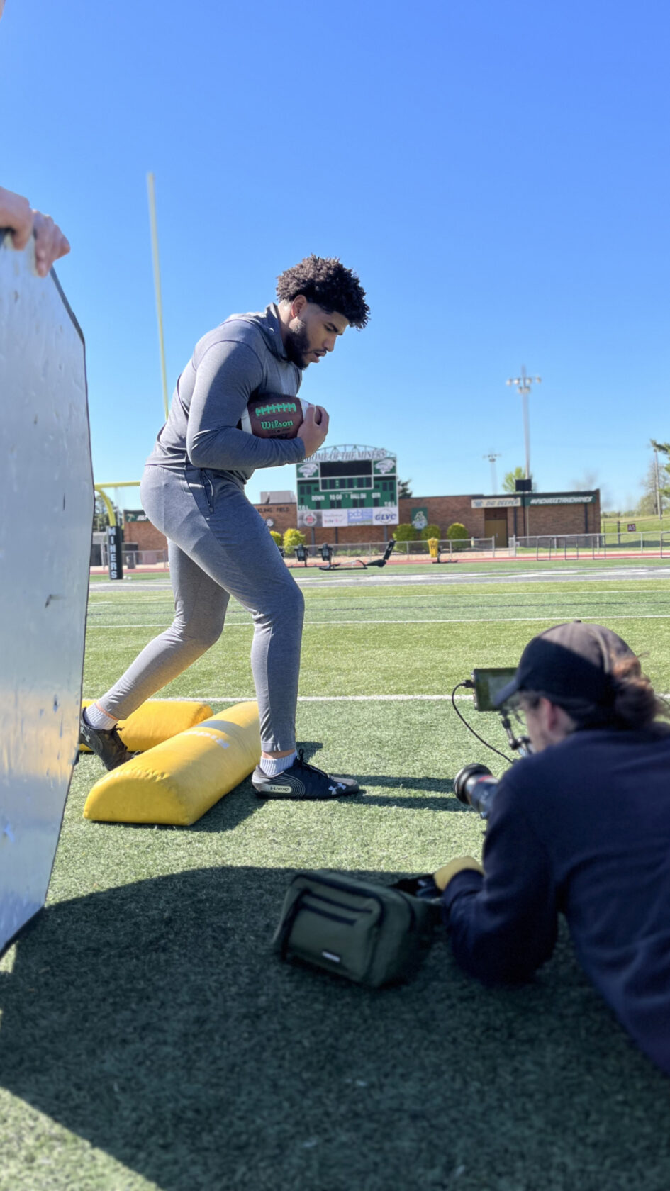 A football player in gray athletic wear runs past yellow tackling dummies on a field as a photographer lies on the ground, capturing the action. A person holds a white reflector to the left. A scoreboard is visible in the background.