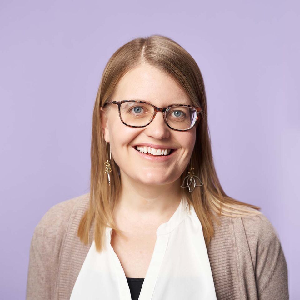A smiling woman with straight, blonde hair wearing glasses, dangling earrings, a white blouse, and a beige cardigan, stands in front of a plain light purple background.