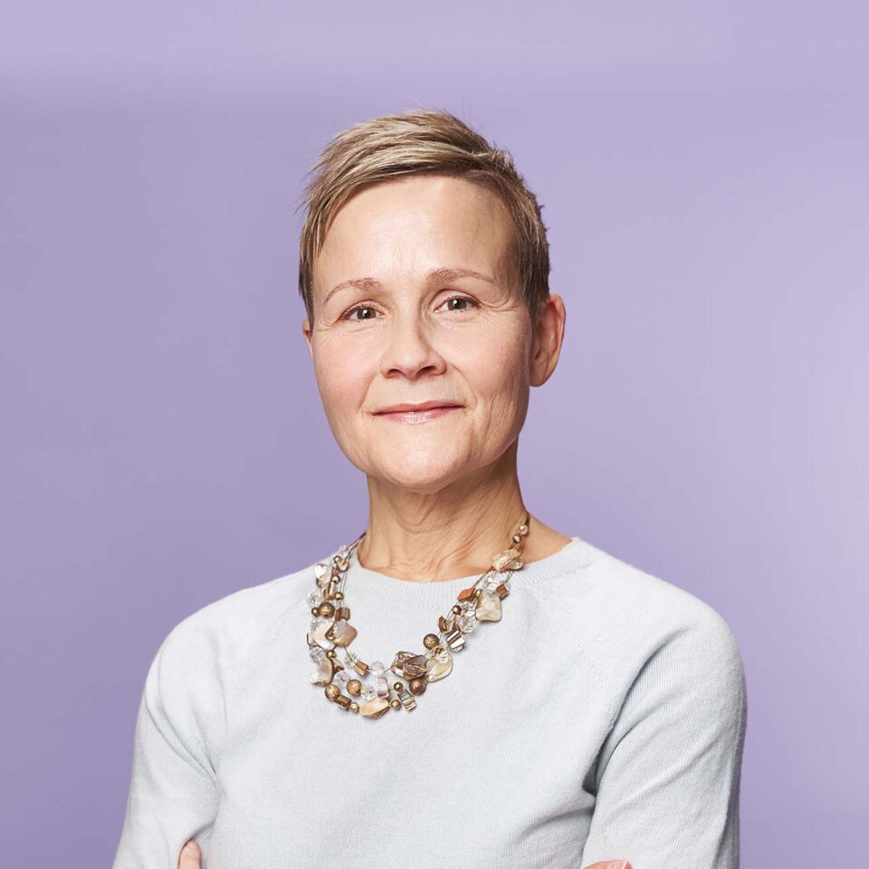 A confident woman with short hair wearing a light-colored top and a beaded necklace stands against a plain lavender background, smiling gently with her arms crossed.