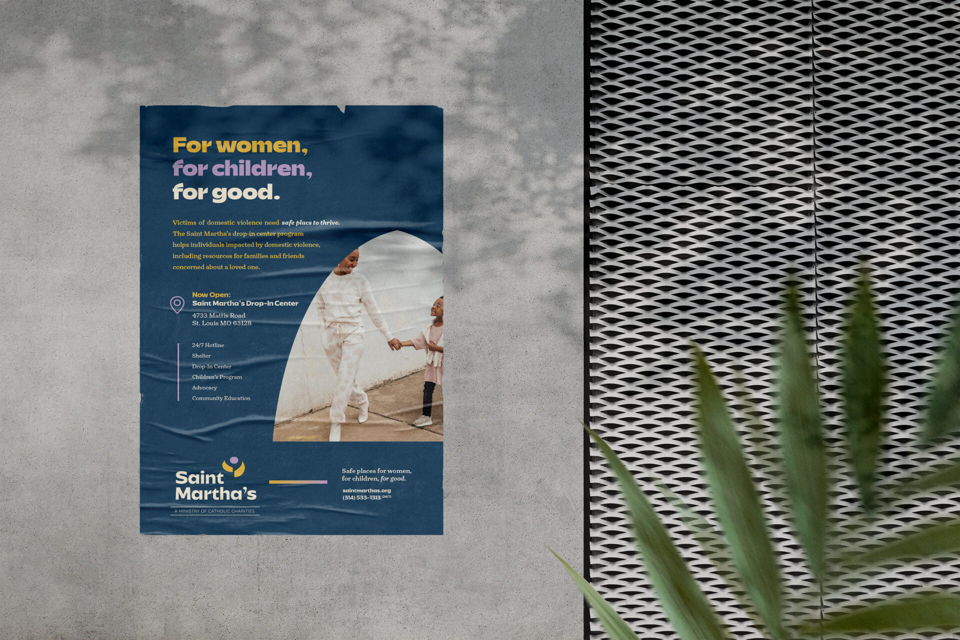 A blue poster on a concrete wall reads “For women, for children, for good.” It features a photo of two people holding hands and promotes Saint Martha’s services for women and children. A green plant is in the foreground.