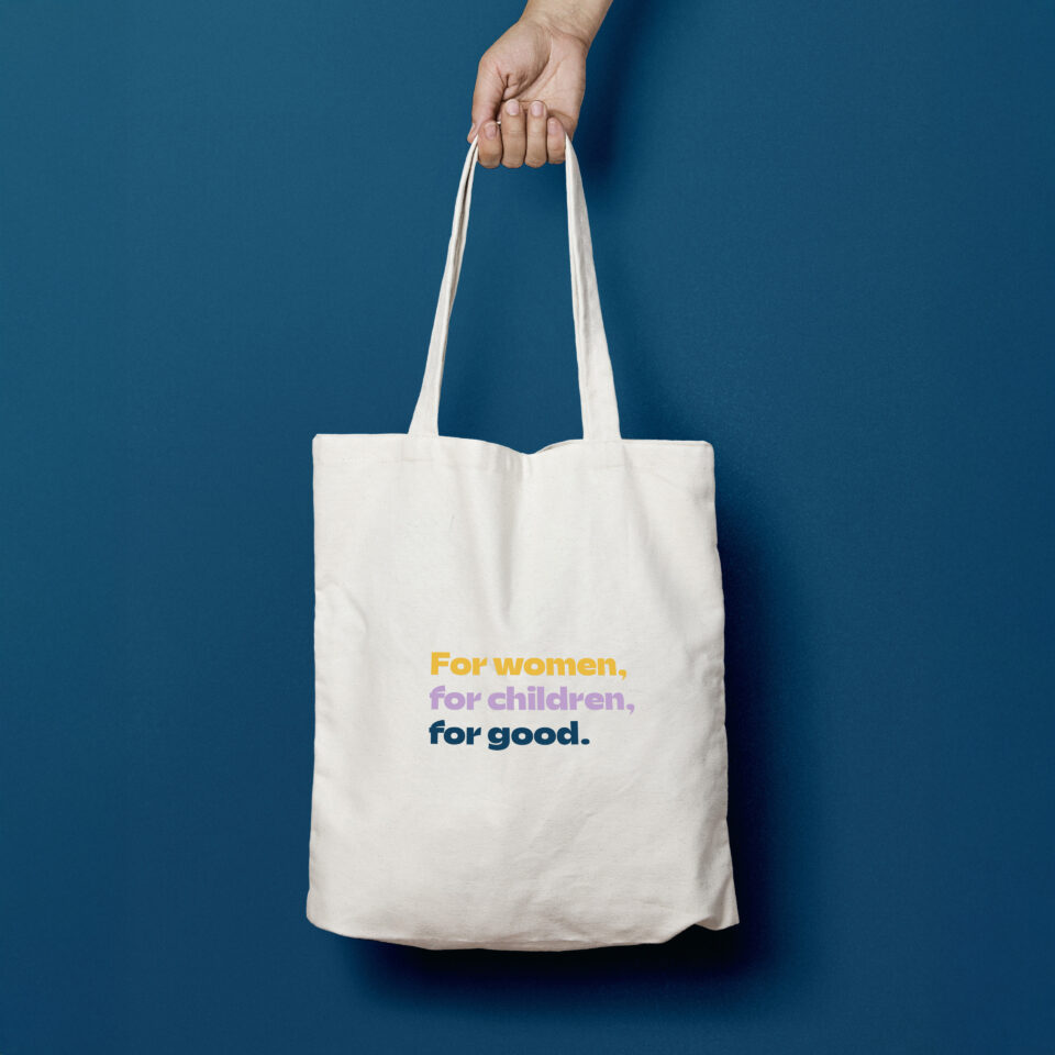 A hand holds a plain white tote bag against a blue background. The bag has text in yellow, purple, and dark blue that reads: For women, for children, for good.