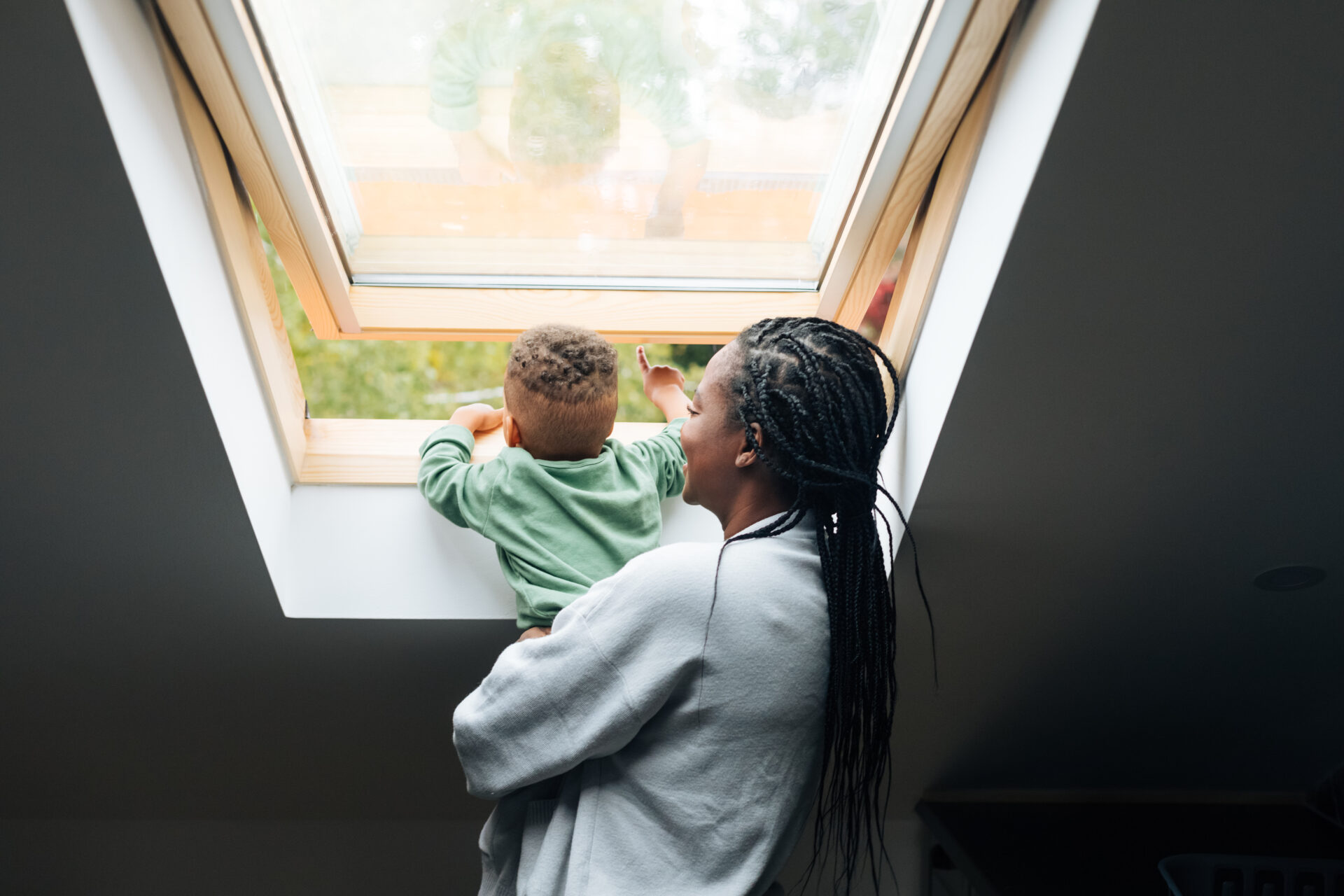 A woman holding a small child looks out through an open skylight window. The child is reaching toward the window, and both appear to be enjoying the view outside. Natural light fills the room.