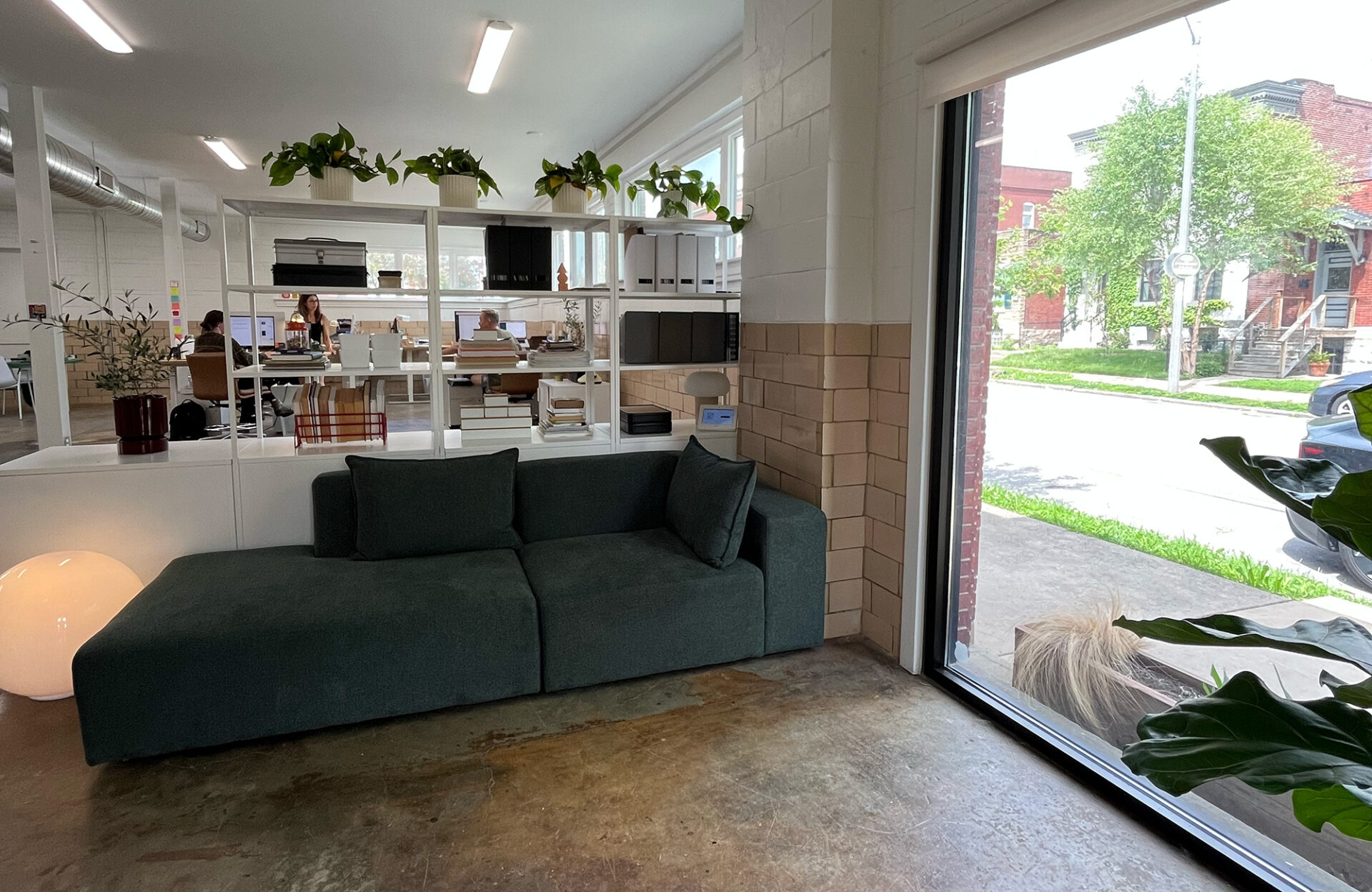Modern office space with a dark green couch near a large window, white shelves with plants and office supplies, and desks with people working in the background. Outside, greenery and buildings are visible.