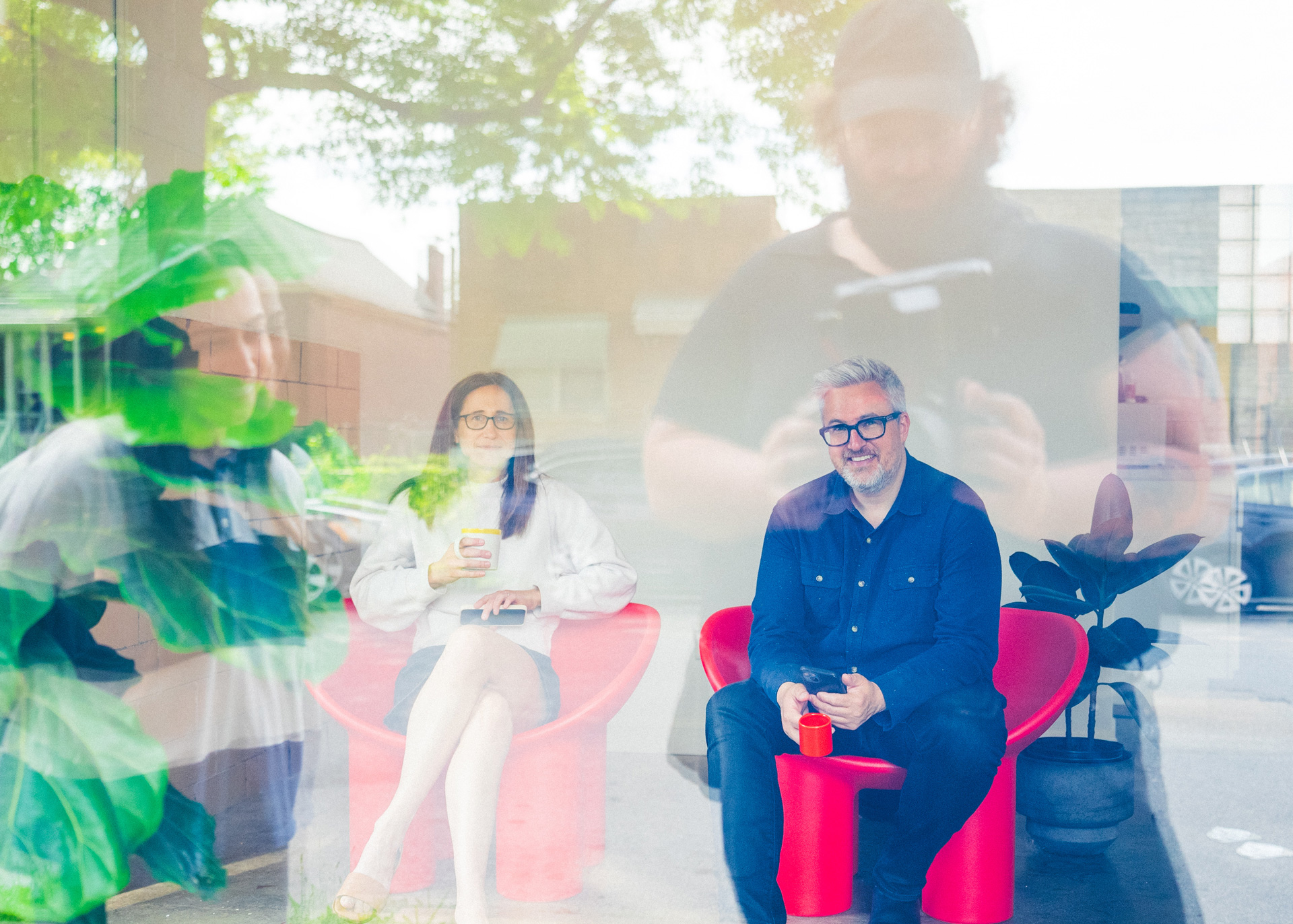 Two people sit on bright red chairs indoors, holding cups and looking outside, while reflections of people and greenery appear in the glass window, blending the interior and exterior scenes.