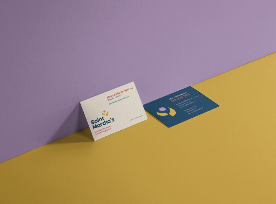 Two business cards for Saint Martha’s are displayed on a yellow surface against a purple wall—one card is white and upright, the other is blue and lying flat, both showing colorful branding and contact details.