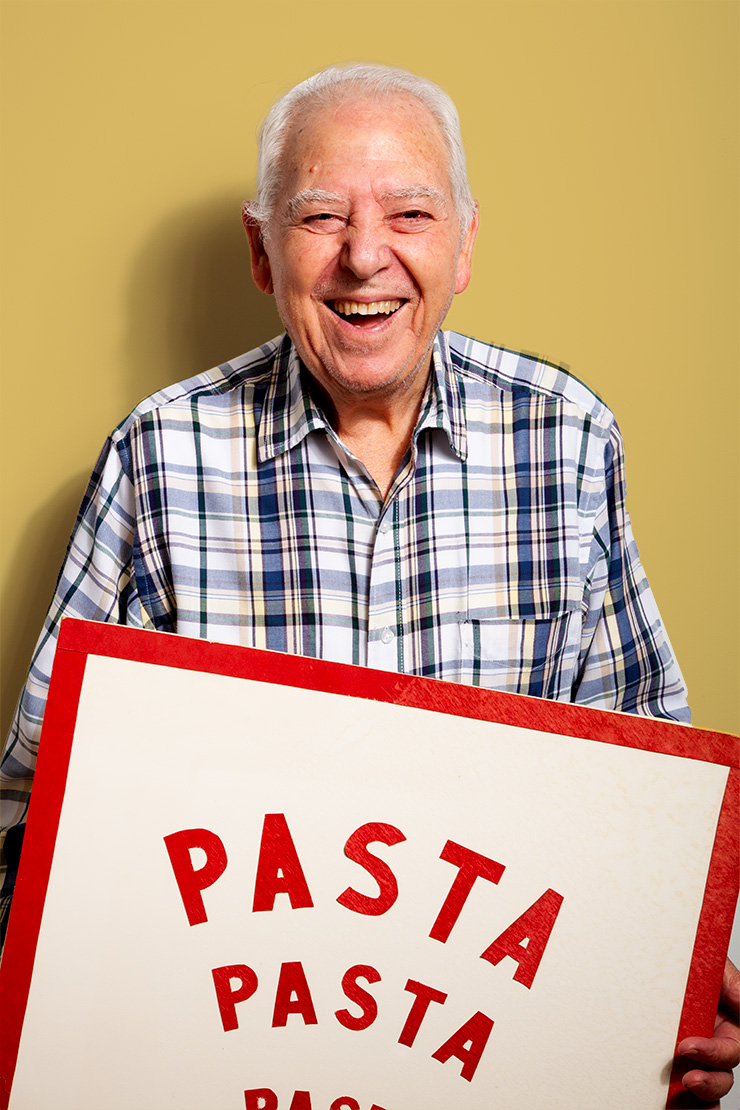 An older man with white hair, wearing a plaid shirt, smiles while holding a large sign that says PASTA in bold red letters, against a plain beige background.