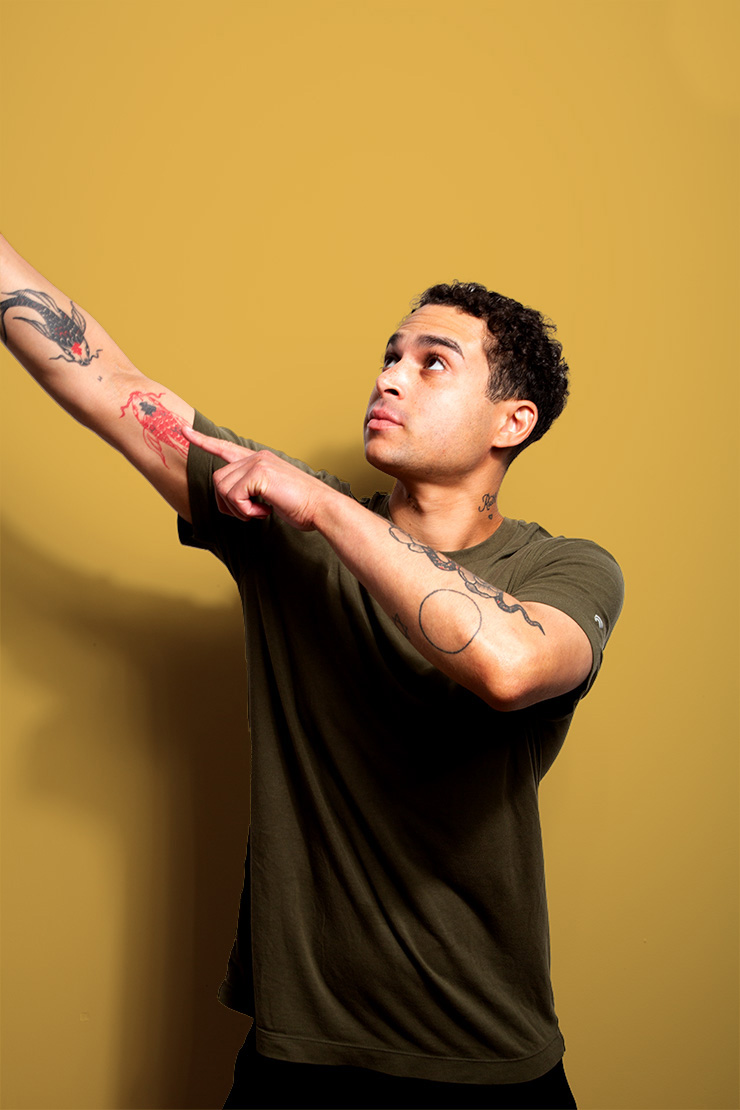A young man with short curly hair and tattoos on both arms poses against a mustard yellow background, pointing upward while looking in the same direction. He wears an olive green t-shirt.