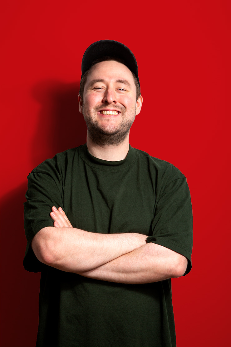 A smiling man wearing a dark green t-shirt and black cap stands with arms crossed in front of a bright red background.