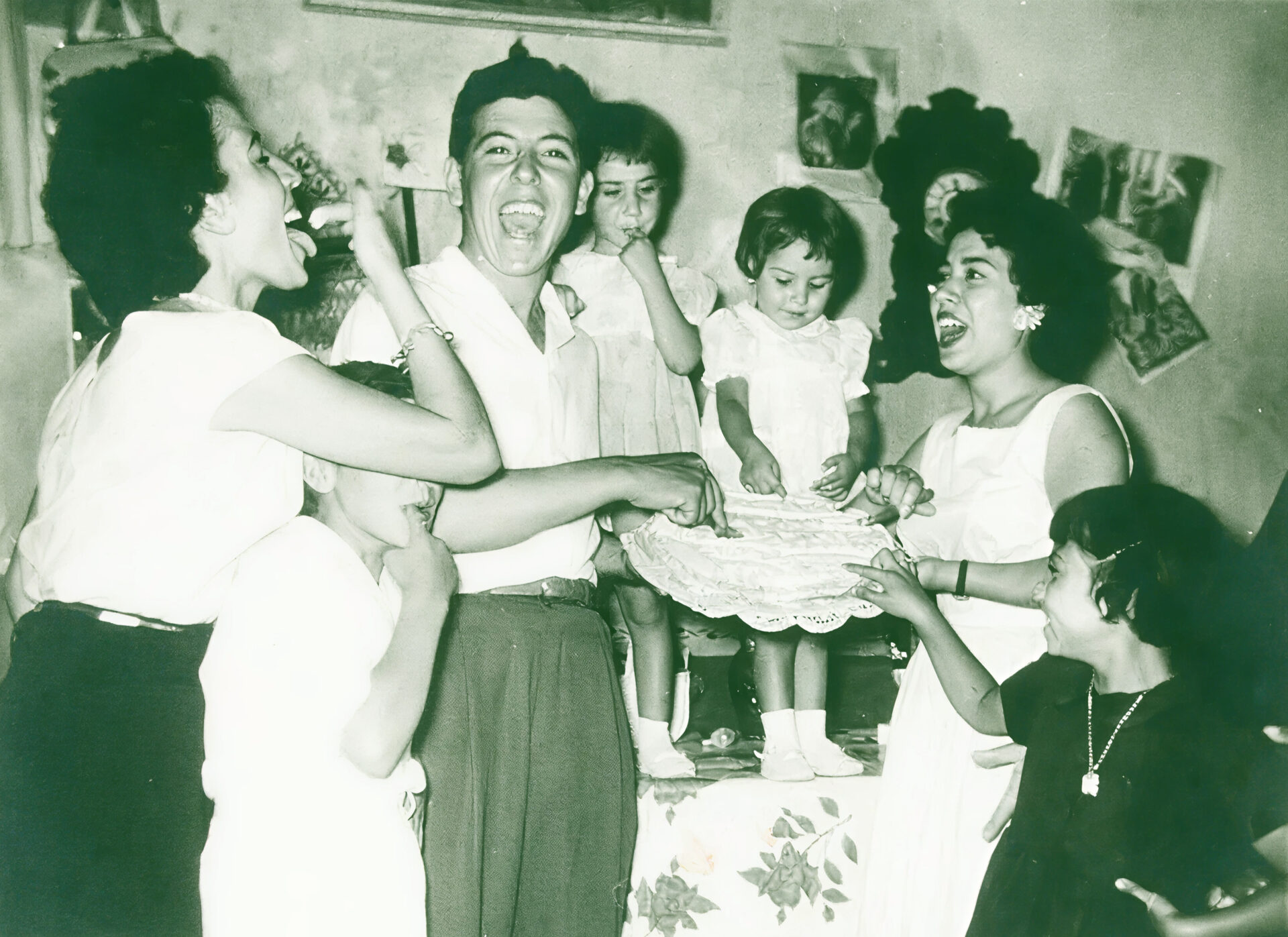 A joyful group of adults and children gather around a table with a cake, laughing and celebrating in a warmly decorated room filled with photos and artwork.