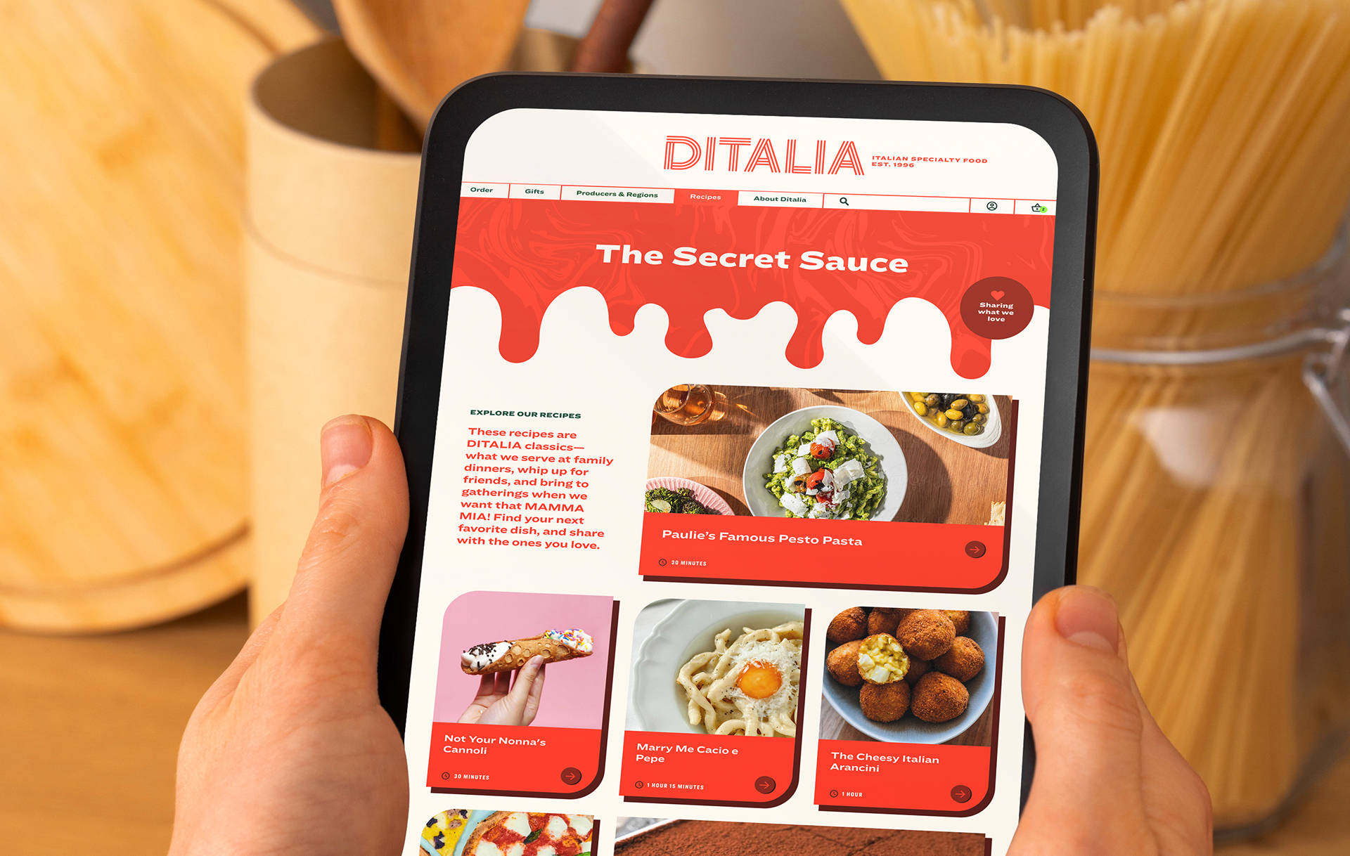 A person holds a tablet displaying the DITALIA website with recipes, including pasta and desserts, on a kitchen counter next to a jar of uncooked spaghetti.