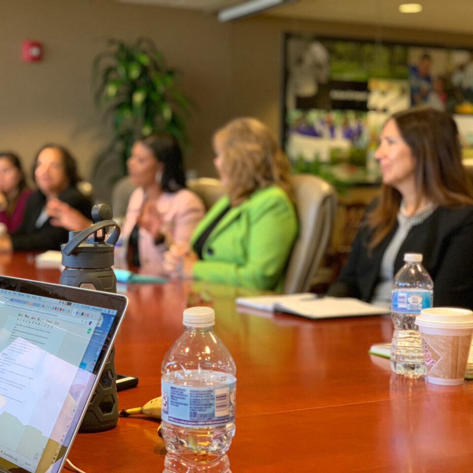 A group of women sit around a conference table having a meeting. A laptop, water bottles, and a coffee cup are on the table. The background is an office with plants and framed photos on the walls.