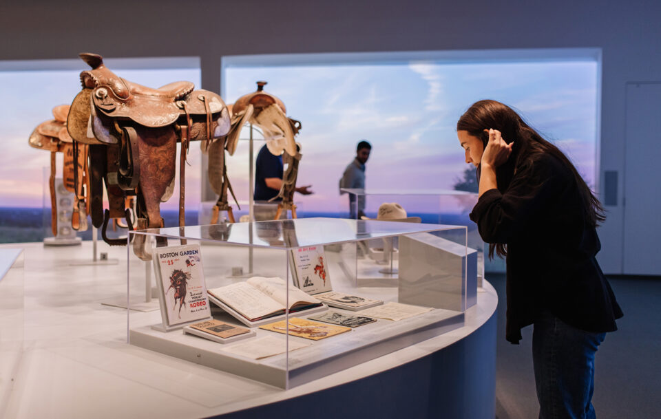 A woman closely examines artifacts and documents displayed in a glass case at a museum, with saddles and other visitors visible in the background against a large window showing a sunset.